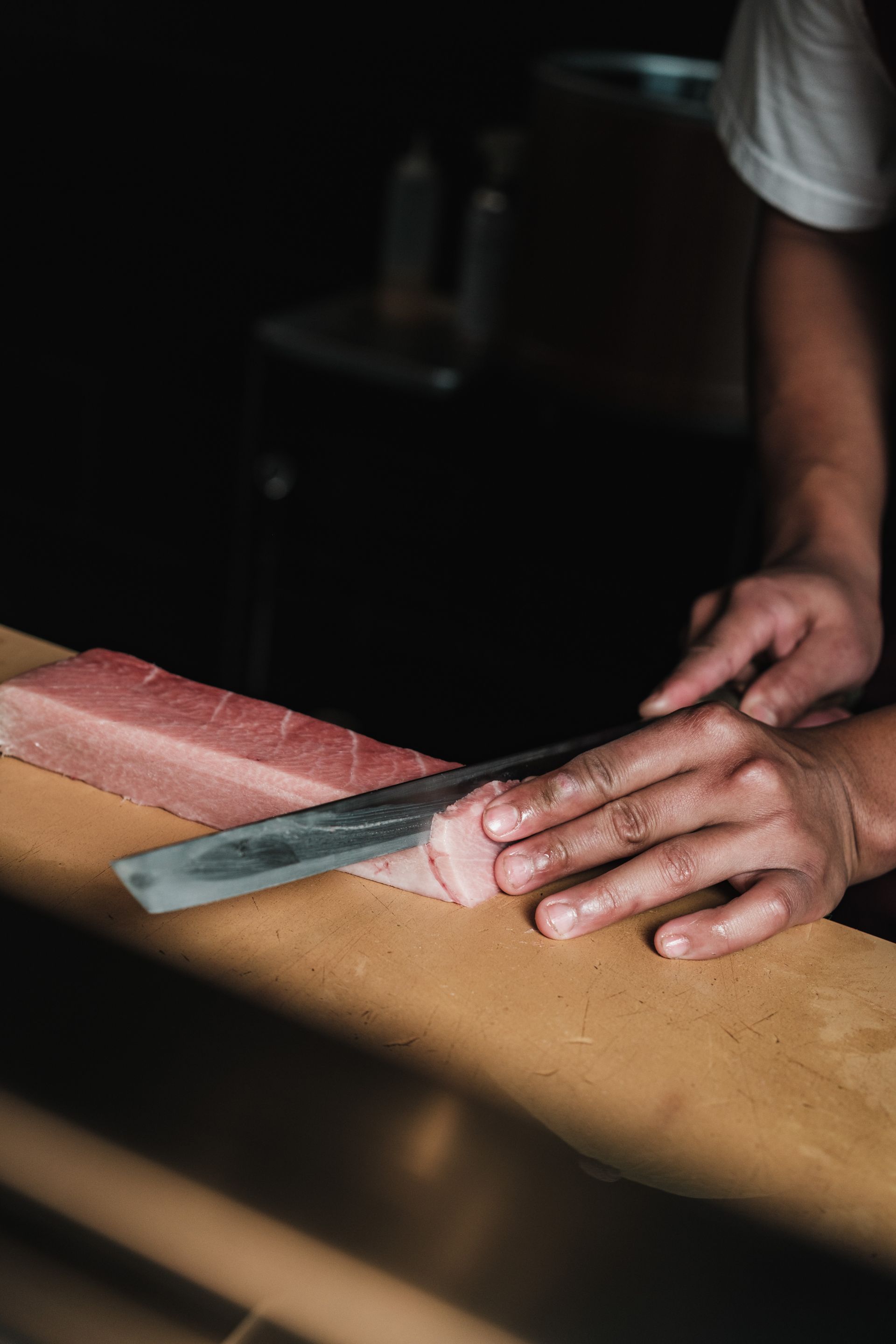 A chef's hands guiding a single-bevel Japanese knife through a block of fresh tuna on a wooden board
