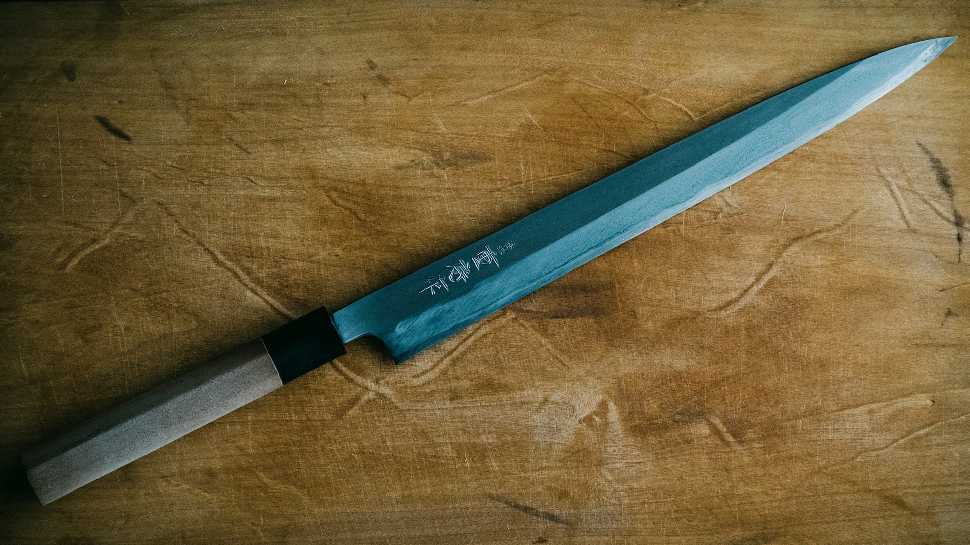 A Japanese yanagiba sashimi knife with kanji engraved on the blade, lying diagonally across a worn wooden board
